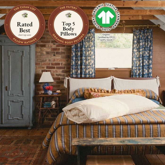 A Circadian Body Pillow in organic cotton lies across the head of a bed in a brick-walled attic bedroom, its zipper open at one end to show the cotton inside, with seals: rated Best by The Clean List, Top 5 organic body pillows on The Filtery, and the GOTS label, the Global Organic Textile Standard.