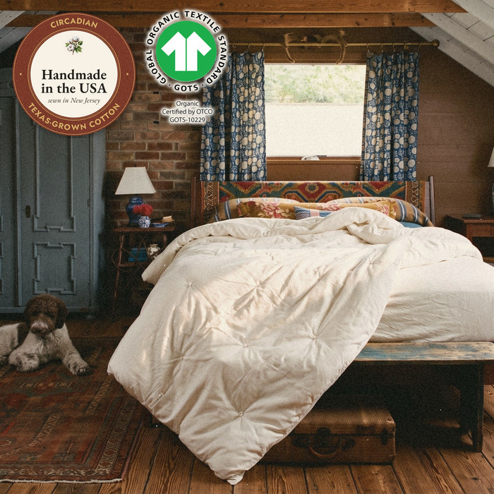 A dog naps on the rug. An Organic Cotton Comforter from Circadian lies thrown back on the bed of a brick attic bedroom, with the Handmade in the USA seal and the GOTS (Global Organic Textile Standard) label, Organic, certified by OTCO, GOTS-10229.