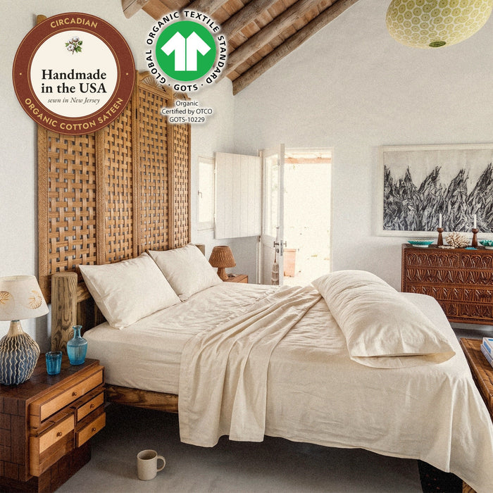A body pillow in a Circadian organic cotton sateen body pillow case along a bed in a beamed bedroom, with seals: handmade in the USA from organic cotton sateen, and the GOTS organic label.