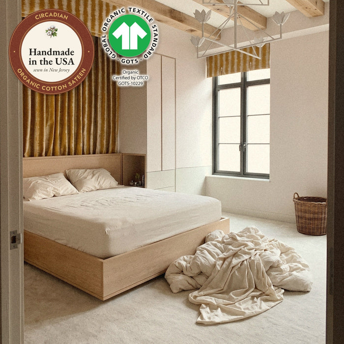 A deep mattress in a Circadian organic cotton sateen fitted sheet, the rest of the bedding in a pile on the floor, with seals: handmade in the USA from organic cotton sateen, and the GOTS organic label.