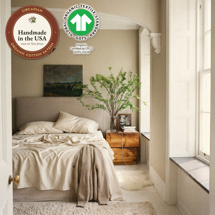 Two pillows in Circadian organic cotton sateen pillowcases on a slept-in bed in a plaster bedroom with branches in a vase, with seals: handmade in the USA from organic cotton sateen, and the GOTS organic label.