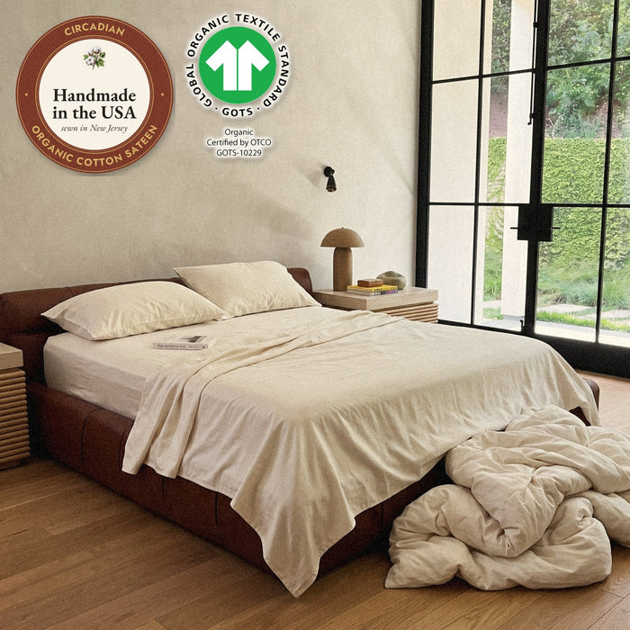 A bed in a Circadian organic cotton sateen sheet set in a plaster bedroom with garden doors, the comforter heaped on the floor, with seals: handmade in the USA from organic cotton sateen, and the GOTS organic label.