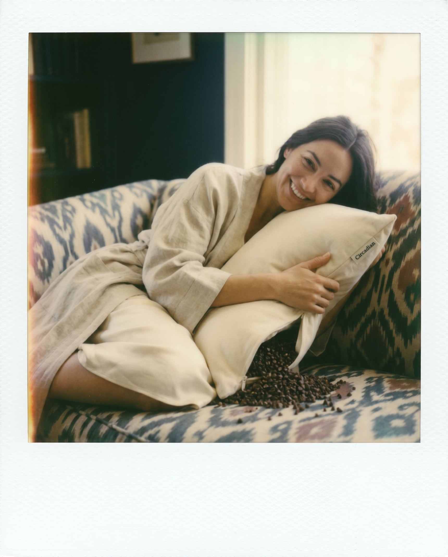Real Circadian customer in linen robe laughing while hugging Buckwheat Pillow, hulls scattered on couch