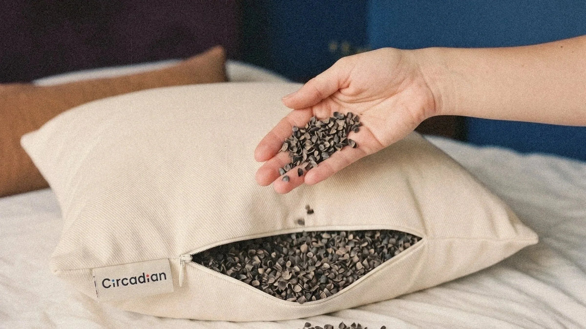 A hand lets buckwheat hulls fall onto an open Circadian Buckwheat Pillow on a bed.