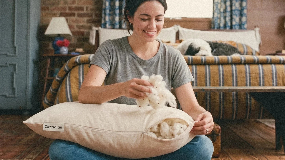 A smiling woman sitting on a rug lifts cotton out of her open Circadian Organic Cotton Pillow.