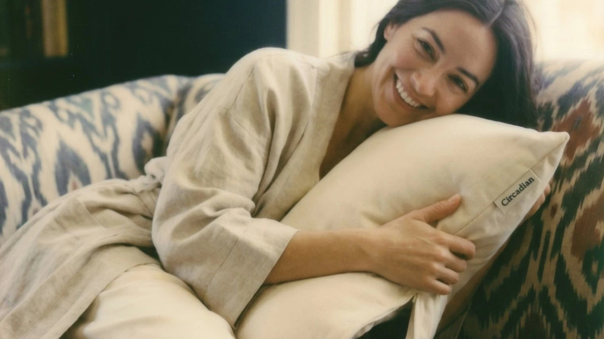 A smiling woman on a sofa hugs her Circadian Buckwheat Pillow, hulls showing at the open zipper.