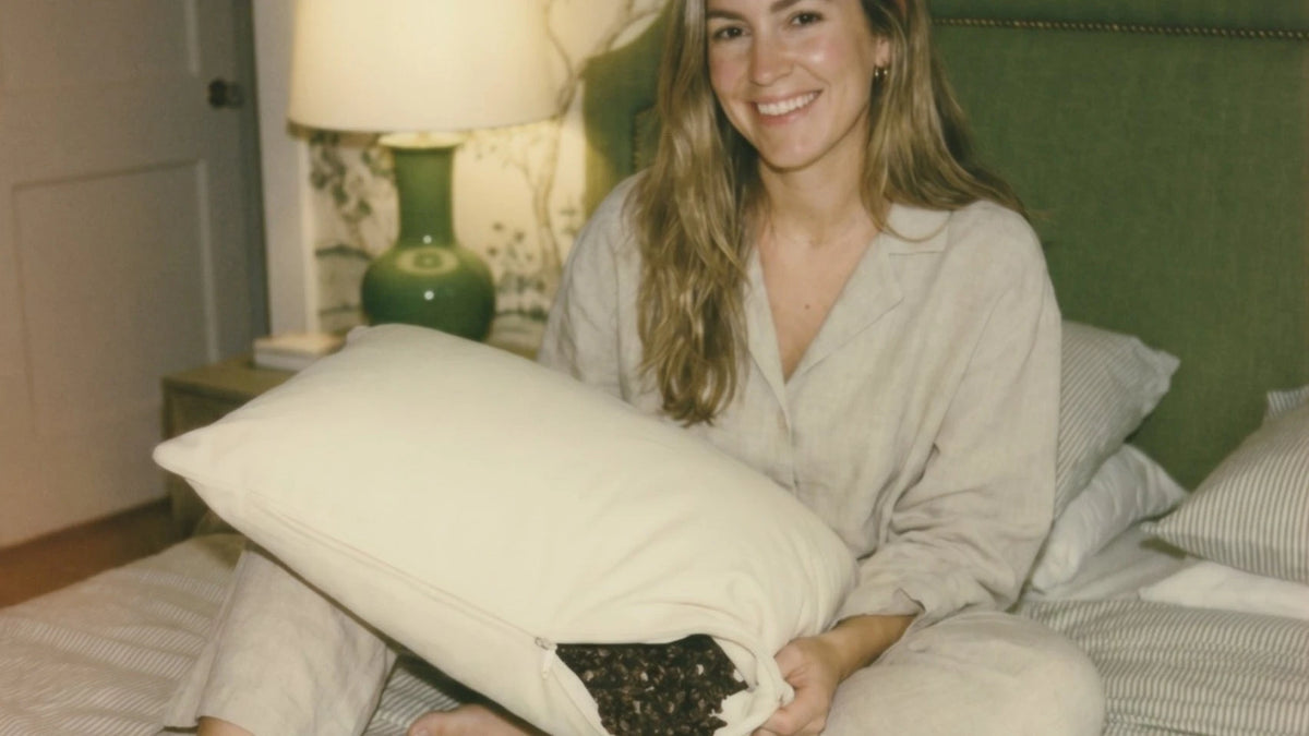 A smiling woman sits on a bed with her open Circadian Buckwheat Pillow, hulls showing.