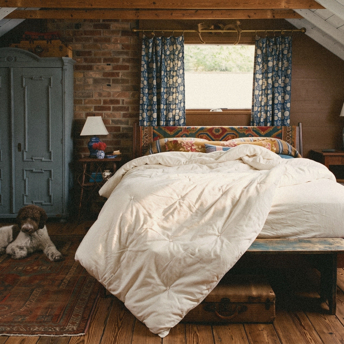 A dog naps on the rug. An Organic Cotton Comforter from Circadian, a hand-tufted duvet insert, lies thrown back on the bed of a brick attic bedroom.