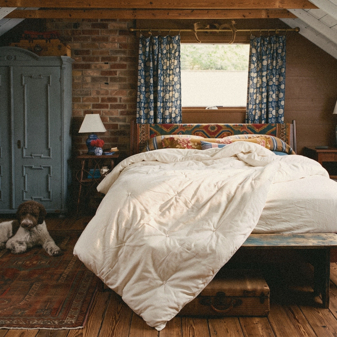 A dog naps on the rug. An Organic Cotton Comforter from Circadian, a hand-tufted duvet insert, lies thrown back on the bed of a brick attic bedroom.
