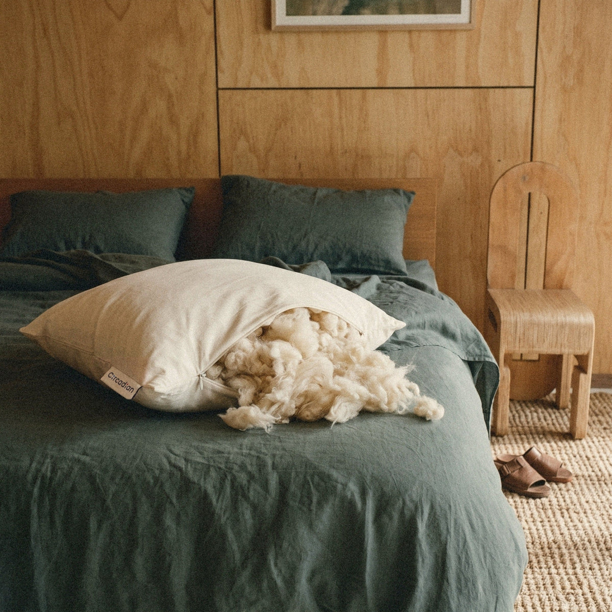 Our Wild-Harvested Kapok Pillow on a green linen bed. Soft, fluffy kapok spills from the open zipper.