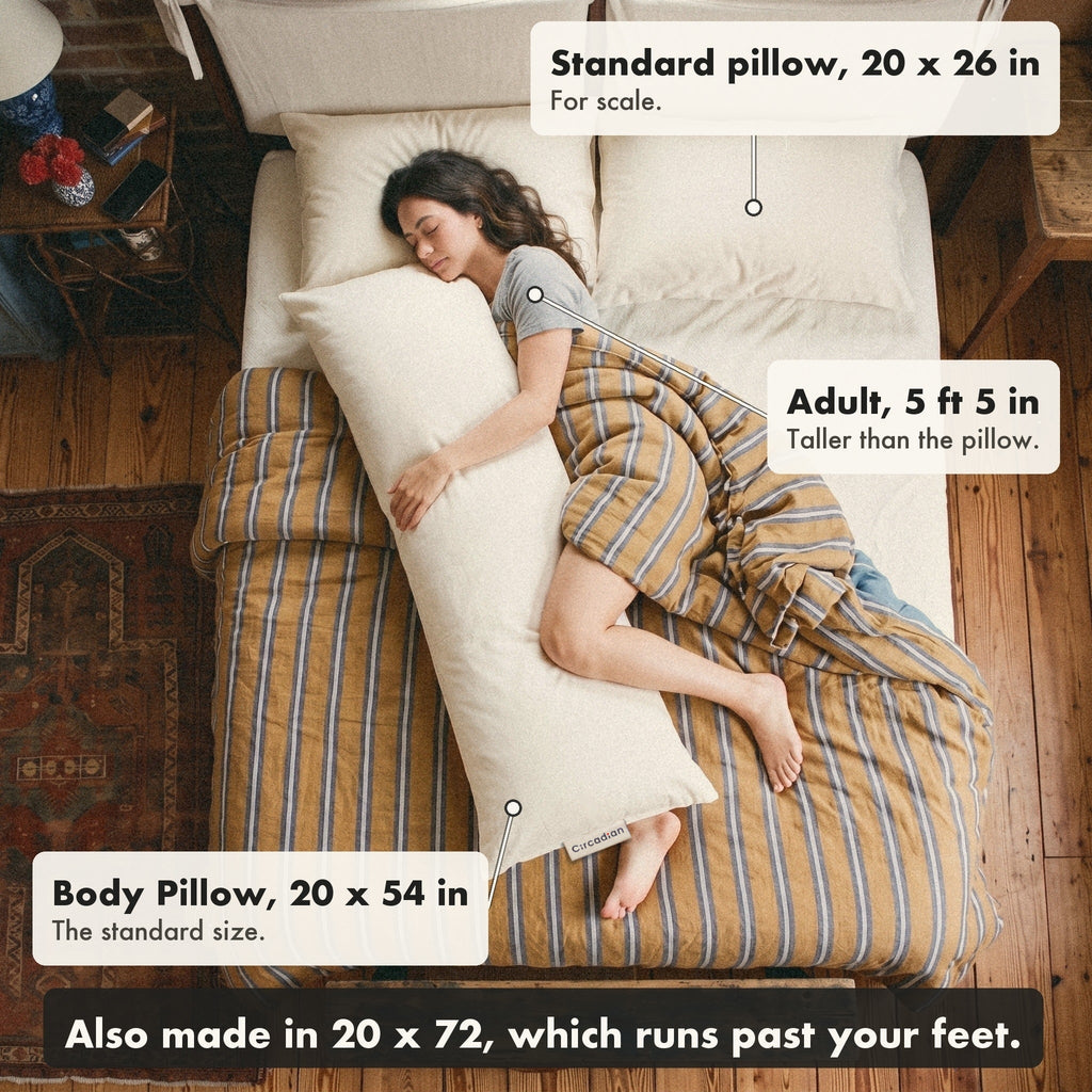Seen from above, a woman of 5 foot 5 sleeps on her side hugging the 20 by 54 inch Circadian Body Pillow on a queen bed, beside a standard 20 by 26 inch pillow; the pillow runs from her chin to her ankles and it also comes in 20 by 72.
