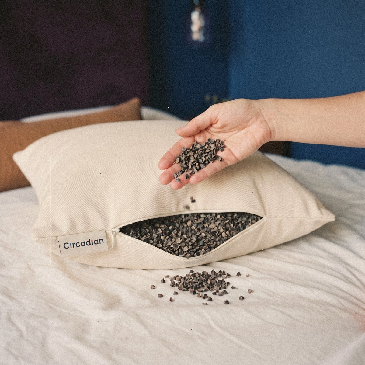 A hand lets a handful of small buckwheat hulls fall back into the open Buckwheat Pillow on a bed.