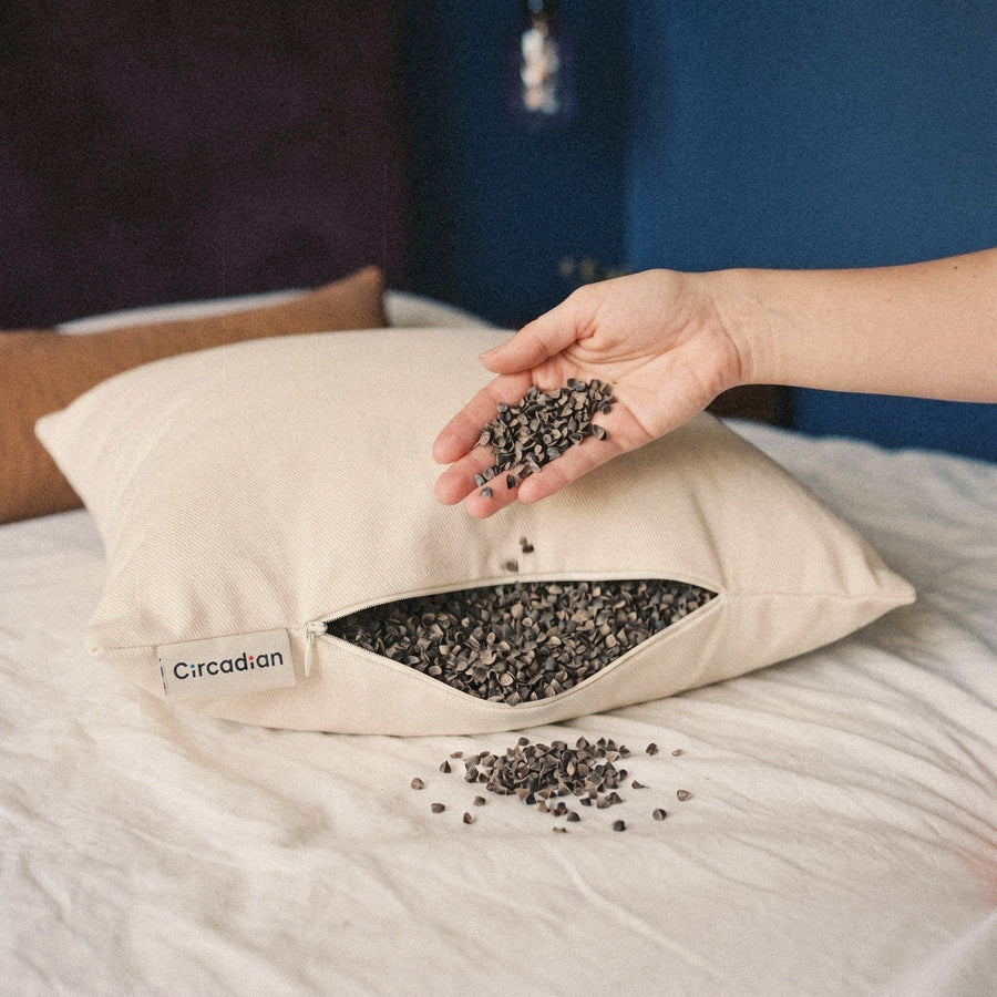 A hand lets a handful of small buckwheat hulls fall back into the open Buckwheat Pillow on a bed.