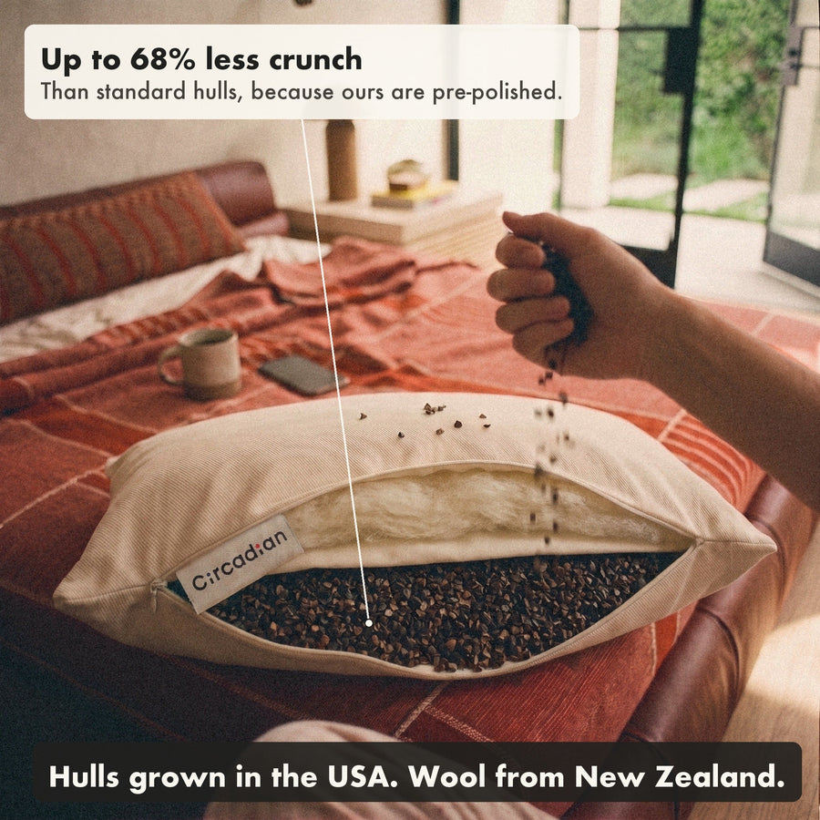 A hand pours buckwheat hulls back into the Buckwool Hybrid Pillow. The labels say up to 68% less crunch than standard hulls, because ours are pre-polished, with hulls grown in the USA and wool from New Zealand.