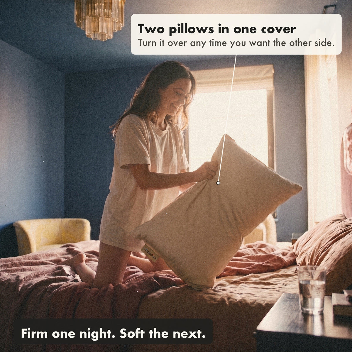 A smiling woman kneeling on her bed turns the Buckwool Hybrid Pillow over, with labels: two pillows in one cover, turn it over any time you want the other side; firm one night, soft the next.