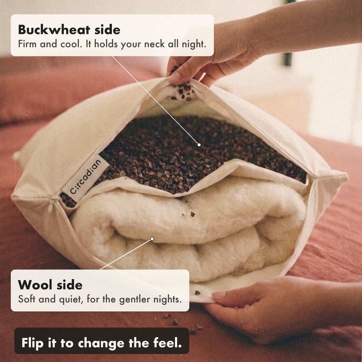 Hands hold the Buckwool Hybrid Pillow open to show buckwheat hulls on one side of a cotton divider and wool on the other, with labels: buckwheat side, firm and cool; wool side, soft and quiet; flip it to change the feel.