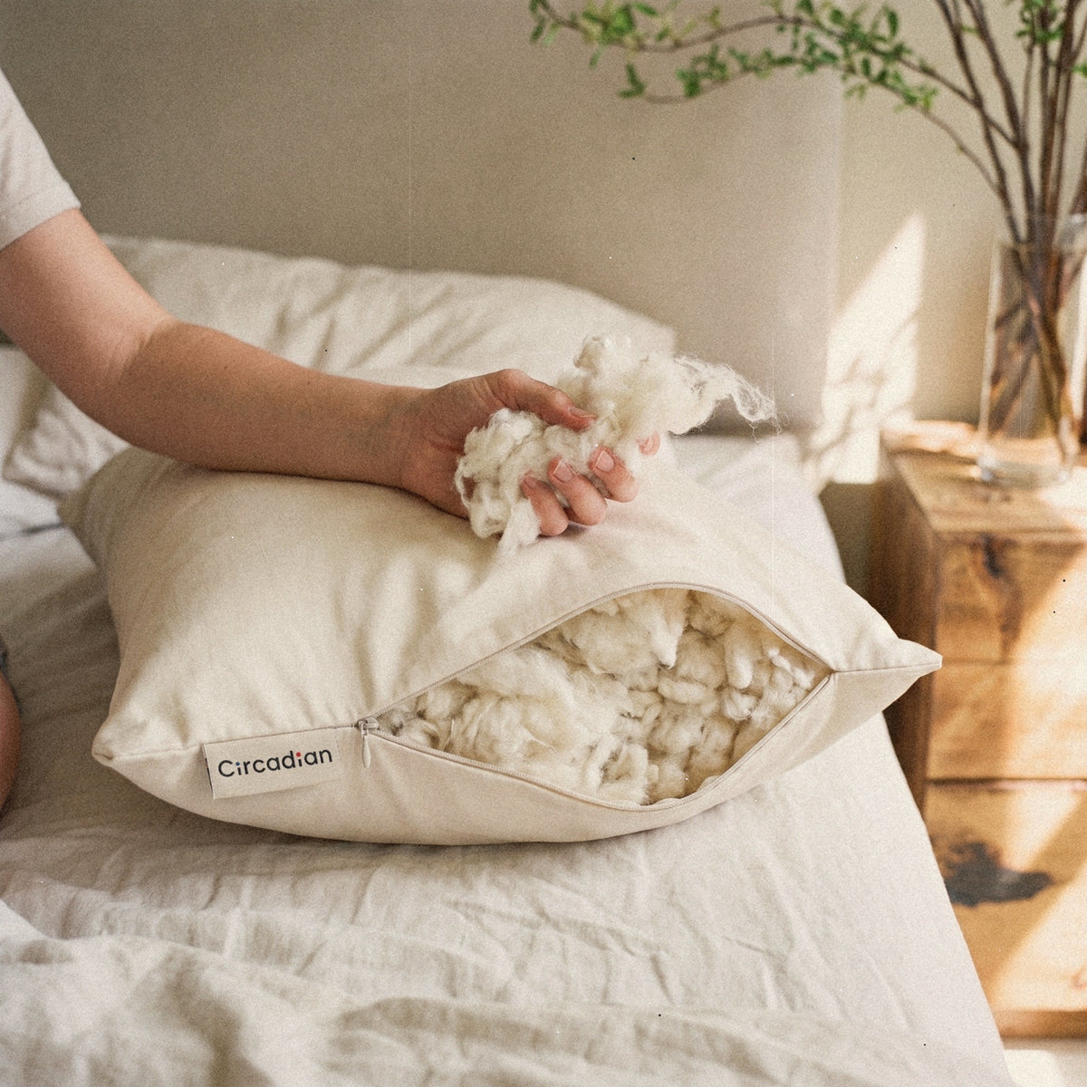 A hand draws a soft tuft of organic cotton out of the open Organic Cotton Pillow on a bed.