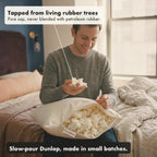 A smiling man sits on his bed with the Tree-Tapped Latex Pillow open on his lap, holding a handful of the soft latex pieces, with labels: tapped from living rubber trees, slow-pour Dunlop made in small batches.