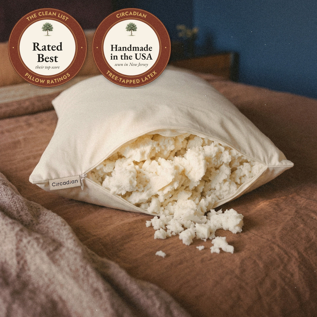A Circadian Tree-Tapped Latex Pillow on a bed with its zipper open and soft, airy pieces of latex spilling out, with seals: rated Best by The Clean List, and handmade in the USA.