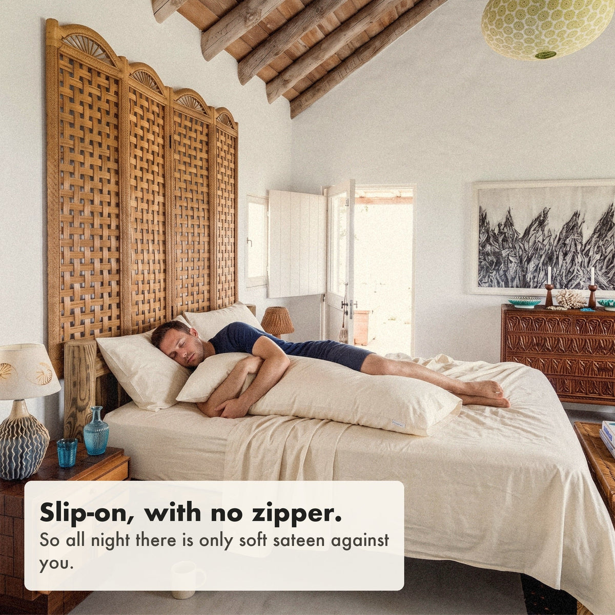 A man asleep on his side hugging a body pillow in an organic cotton sateen case, labelled: slip-on, with no zipper, so all night there is only soft sateen against you.
