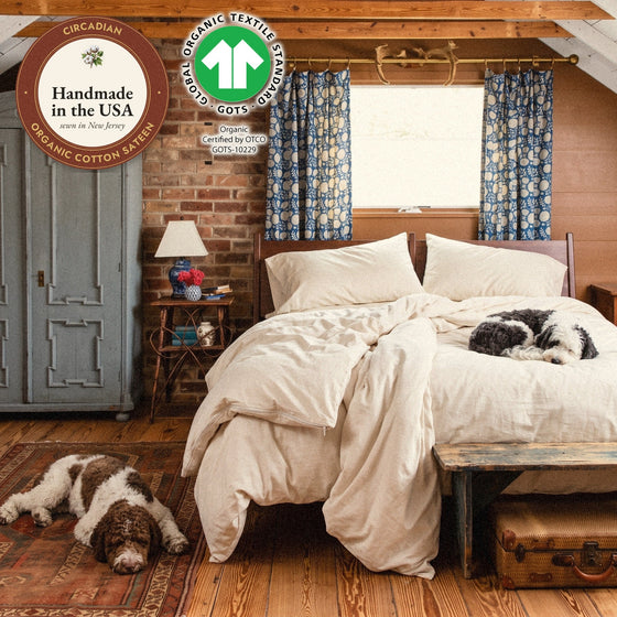 A comforter in a Circadian organic cotton sateen duvet cover on a bed in a brick attic bedroom, two dogs nearby, with seals: handmade in the USA from organic cotton sateen, and the GOTS organic label.