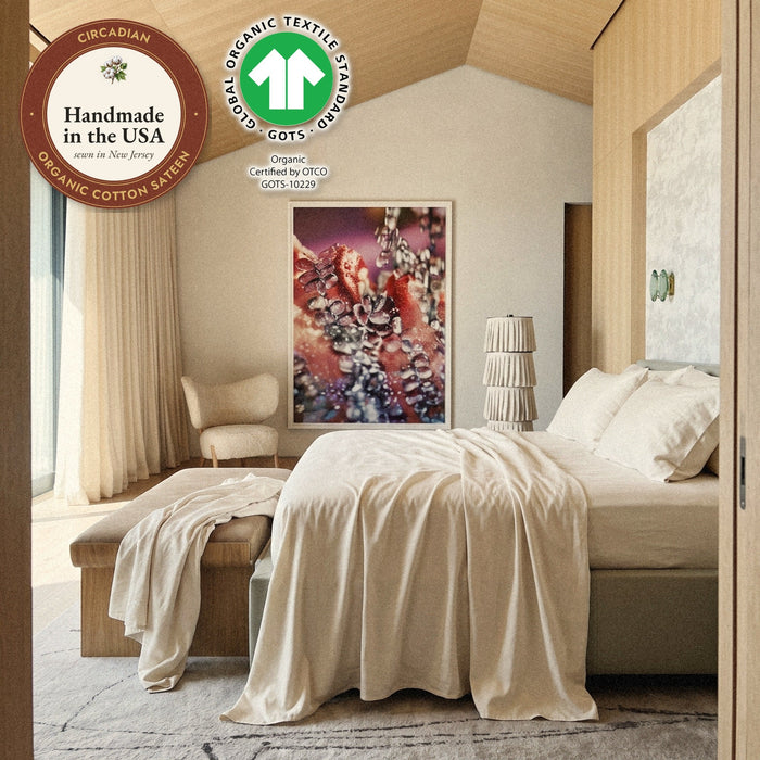 A Circadian organic cotton sateen flat sheet turned back over a bed under a wood ceiling, with seals: handmade in the USA from organic cotton sateen, and the GOTS organic label.