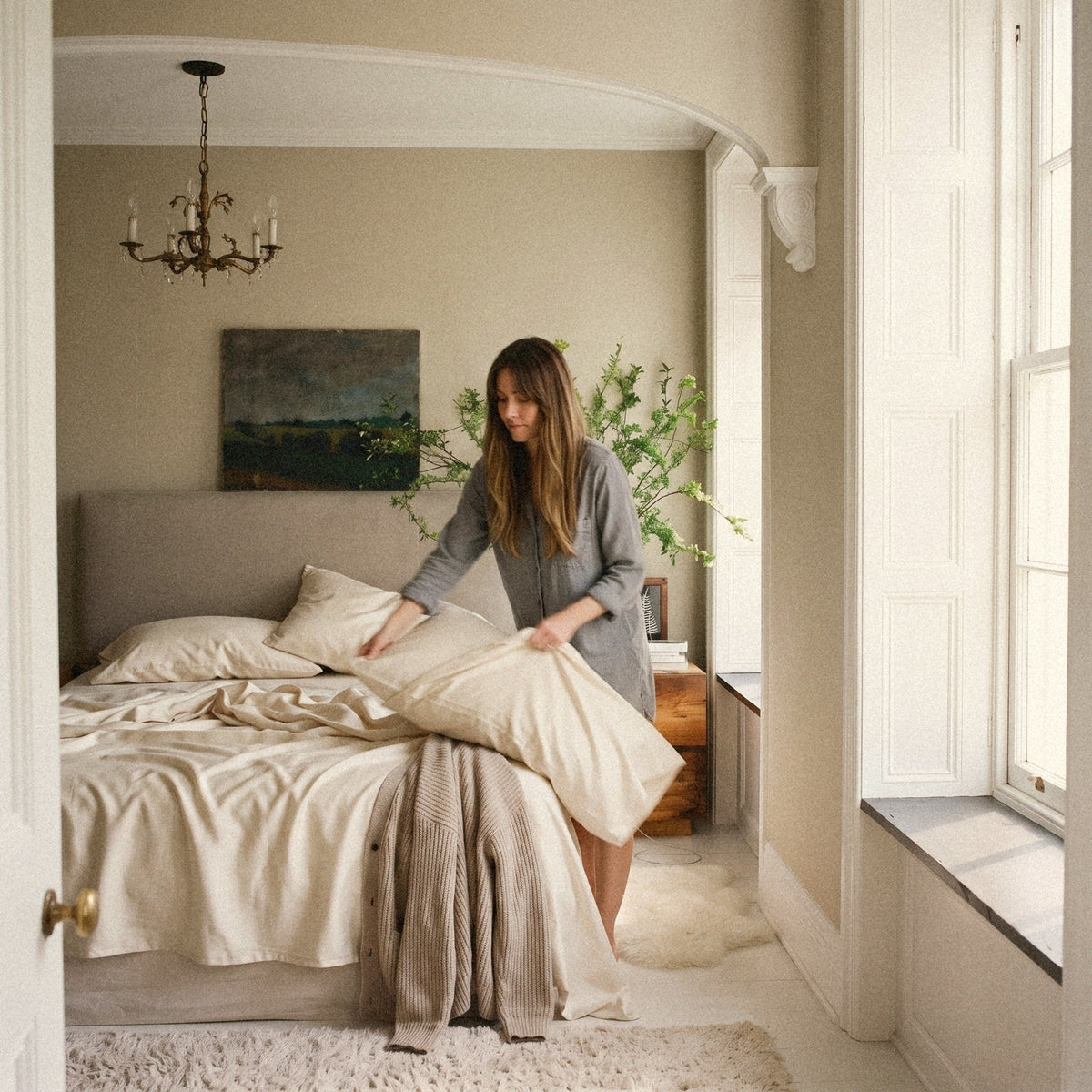 A woman standing by her bed slides a pillow into a Circadian organic cotton sateen pillowcase.
