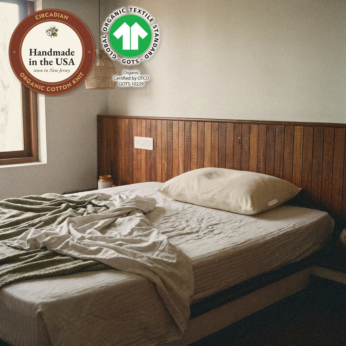 A pillow in the Circadian waterproof organic cotton pillow protector on a bed in a wood-panelled bedroom, with seals: handmade in the USA from organic cotton knit, and the GOTS organic label.