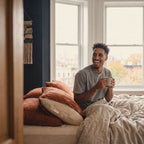 A man sitting up in bed with a mug of coffee, the waterproof pillow protector showing at the end of his pillowcase.