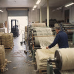 A mill worker guides a thin, airy web of undyed wool as it comes off a carding machine and gathers into soft batting.