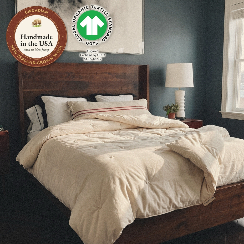 Morning sun on a dark wood bed. The Circadian Organic Wool Comforter, a hand-tufted duvet insert, lies thrown back, with the Handmade in the USA seal and the label of GOTS, the Global Organic Textile Standard (Organic, certified by OTCO, GOTS-10229).