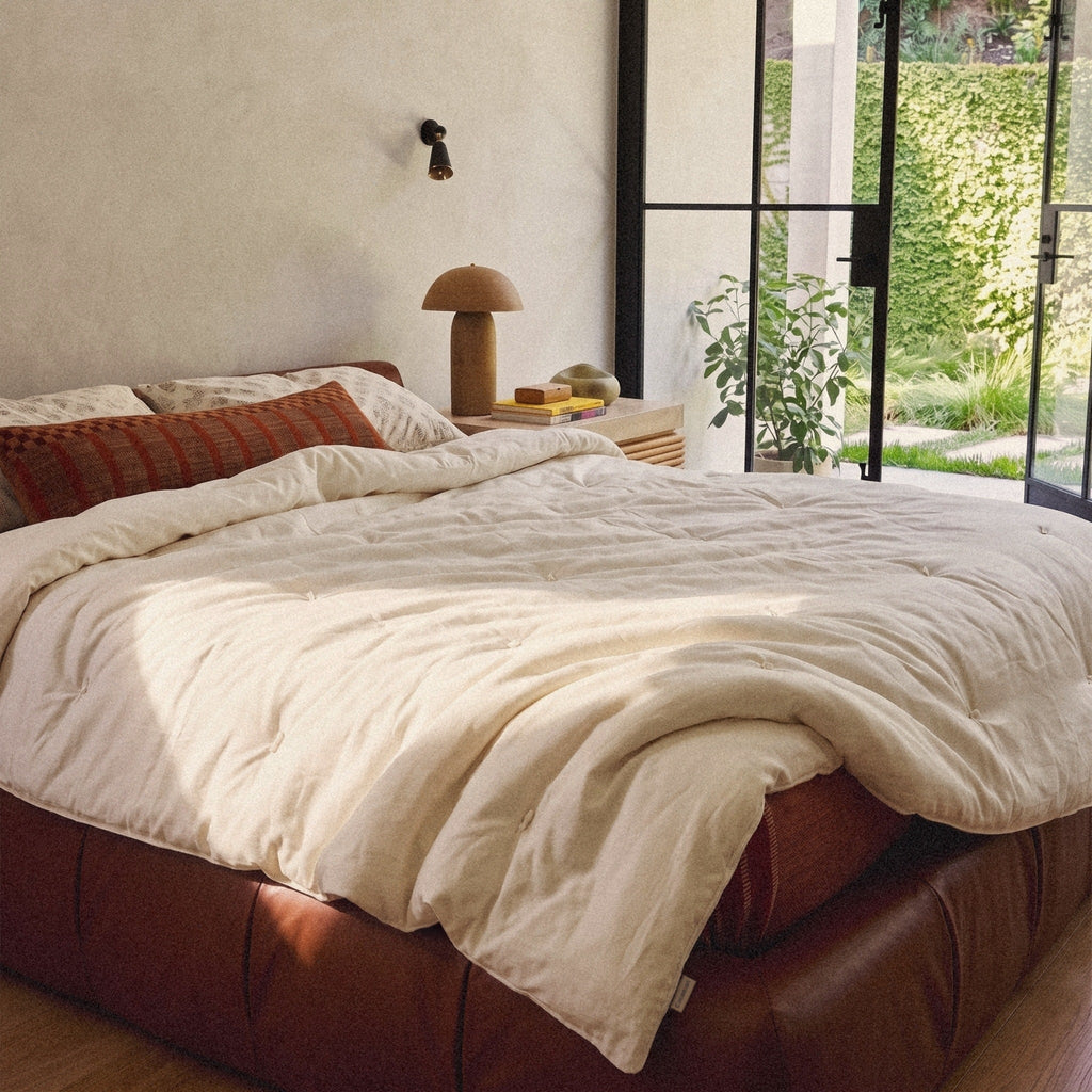 Our Organic Wool Comforter thrown back on a leather bed in a bright plaster bedroom with doors open to the garden, its woven Circadian label at the edge.
