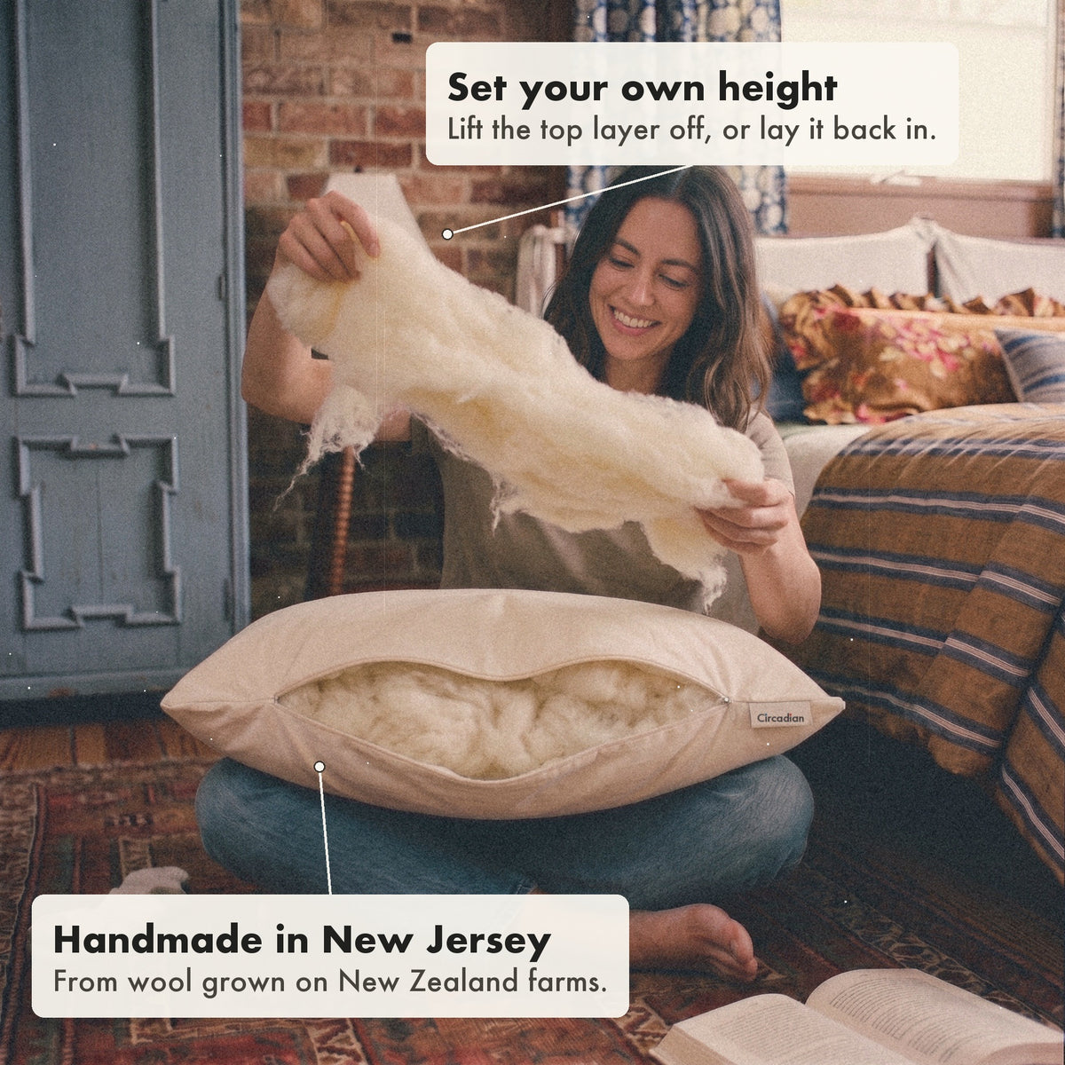 A smiling woman sitting on a rug lifts a soft layer of wool out of the Organic Wool Pillow to set its height, with labels: set your own height, handmade in New Jersey from wool grown on New Zealand farms.